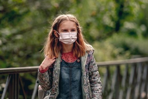 Pretty school girl wearing face mask standing in autumn park and looking at camera. Beautiful female kid after school in pandemic coronavirus covid time