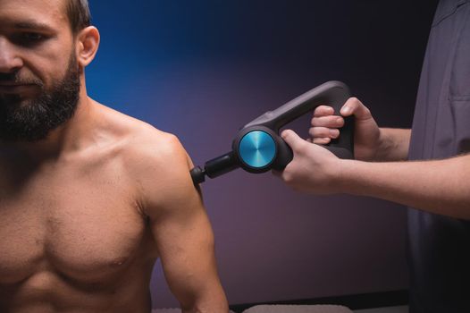 Shoulder massage with a percussion massager in a professional massage parlour. Working as a percussion massager in a dark room.
