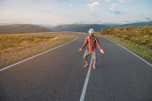 Smiling bearded friendly Caucasian man in a shirt and shorts with a backpack greets you spreading his arms to the sides and sitting down. while standing on a country asphalt road high in the mountains at sunset..