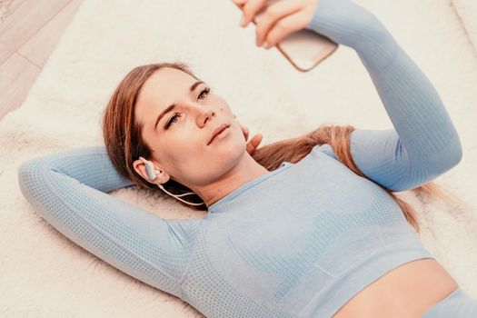 Top view portrait of relaxed woman listening to music with headphones lying on carpet at home. She is dressed in a blue tracksuit