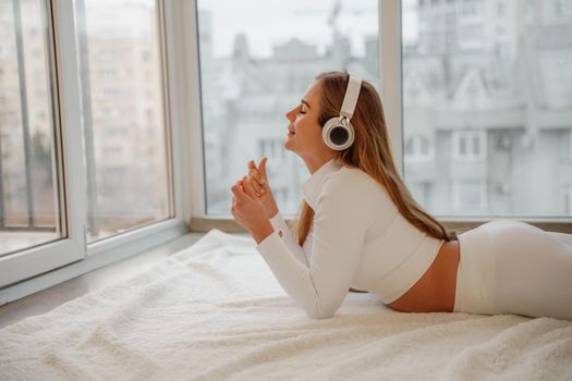 Side view portrait of relaxed woman listening to music with headphones lying on carpet at home. She is dressed in a white tracksuit