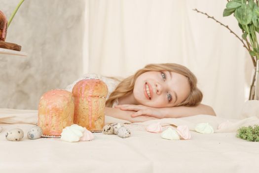 A girl with long hair in a light dress is sitting at the Easter table with cakes, spring flowers and quail eggs. Happy Easter celebration