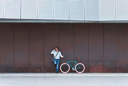 Young male with sunglasses and shoulder bag standing nest to a fixed gear bike while calling by phone