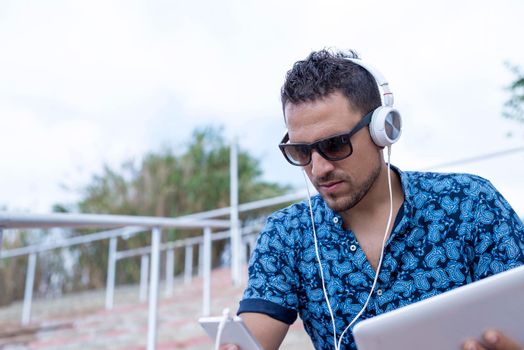 Young male with headphones and sunglasses sitting on stairs outdoors using tablet