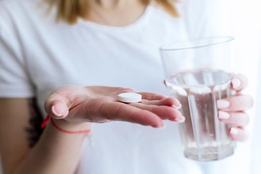 A young woman holds a glass of water and a pill for pain in the head or abdomen.