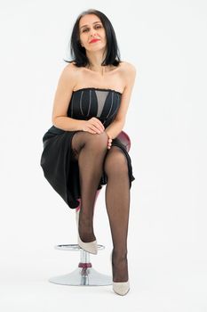 Middle aged woman starting getting grey-haired is posing in studio while sitting on the chair in black dress and stockings on white background, middle age skincare cosmetics, cosmetology concept.