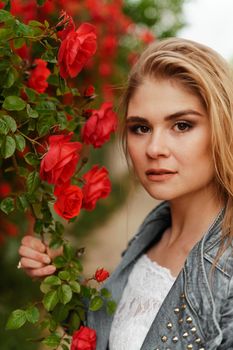 A cute blonde stands near a bush with red roses. Attractive European woman in a denim jacket