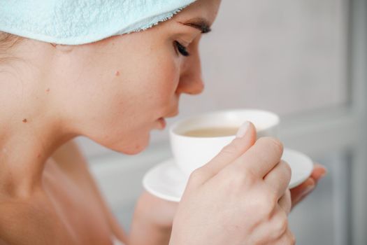 Young serene relaxed woman in spa bath towel drinking hot beverage tea coffee after taking shower bath at home. Beauty treatment, hydration concept