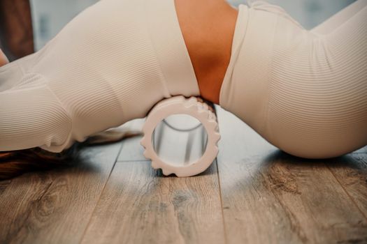 Adult athletic woman, in white bodysuit, performing fascia exercises on the floor - caucasian woman using a massage foam roller - a tool to relieve tension in the back and relieve muscle pain - the concept of physiotherapy and stretching training.