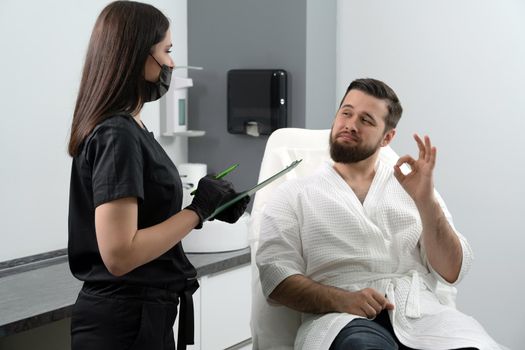 Expert female cosmetologist giving consultation to young man patient sitting on couch in beautician office of aesthetic clinic