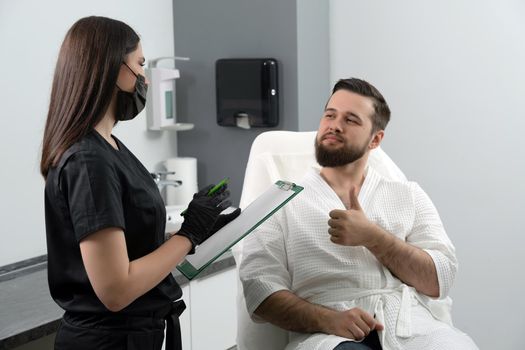 Expert female cosmetologist giving consultation to young man patient sitting on couch in beautician office of aesthetic clinic