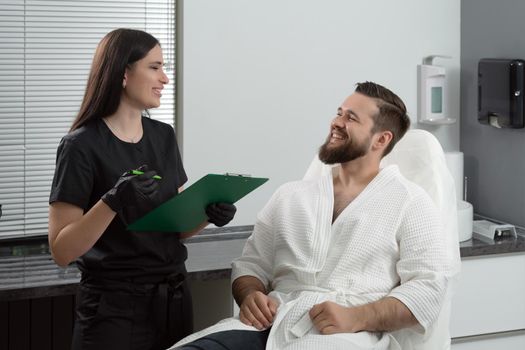 Expert female cosmetologist giving consultation to young man patient sitting on couch in beautician office of aesthetic clinic