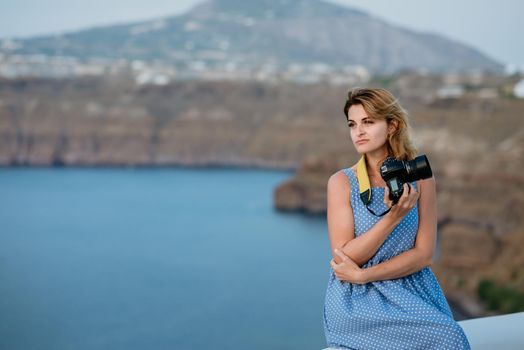 Woman in a blue dress sitting on the rocks on the beach and taking pictures with a SLR camera. Sea and mountains in the background