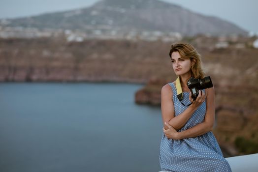 Woman in a blue dress sitting on the rocks on the beach and taking pictures with a SLR camera. Sea and mountains in the background