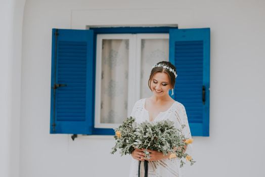 Handsome beautiful caucasian bride posing near white wall with blue windows. Young attractive bride with the bouquet of flowers.