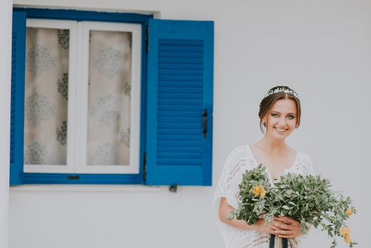 Handsome beautiful caucasian bride posing near white wall with blue windows. Young attractive bride with the bouquet of flowers.