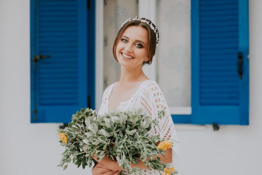 Handsome beautiful caucasian bride posing near white wall with blue windows. Young attractive bride with the bouquet of flowers.