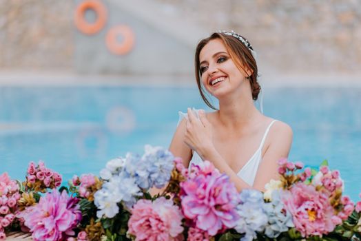 Beautiful bride in a beautiful white swimsuit and garlands of flowers, standing in swimming pool with flower on turquoise water, beautiful girl in water pool in sunny day.