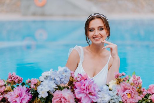 Beautiful bride in a beautiful white swimsuit and garlands of flowers, standing in swimming pool with flower on turquoise water, beautiful girl in water pool in sunny day.