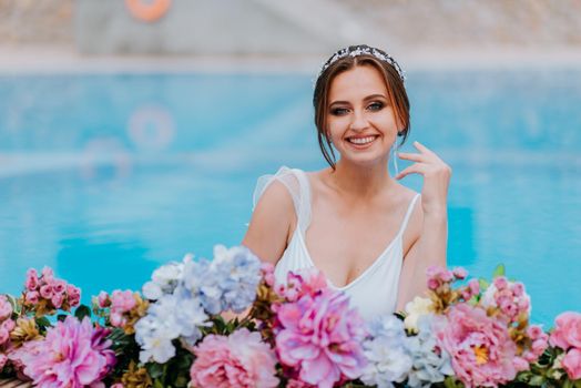 Beautiful bride in a beautiful white swimsuit and garlands of flowers, standing in swimming pool with flower on turquoise water, beautiful girl in water pool in sunny day.