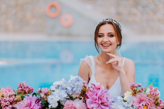 Beautiful bride in a beautiful white swimsuit and garlands of flowers, standing in swimming pool with flower on turquoise water, beautiful girl in water pool in sunny day.