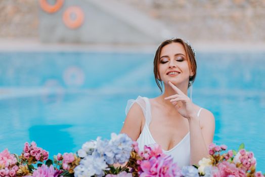 Beautiful bride in a beautiful white swimsuit and garlands of flowers, standing in swimming pool with flower on turquoise water, beautiful girl in water pool in sunny day.