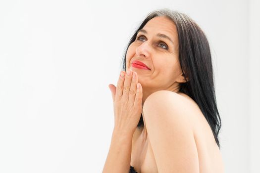 Middle aged woman starting getting grey-haired is posing in studio with naked shoulders on white background, face skin care beauty, middle age skincare cosmetics, cosmetology concept.
