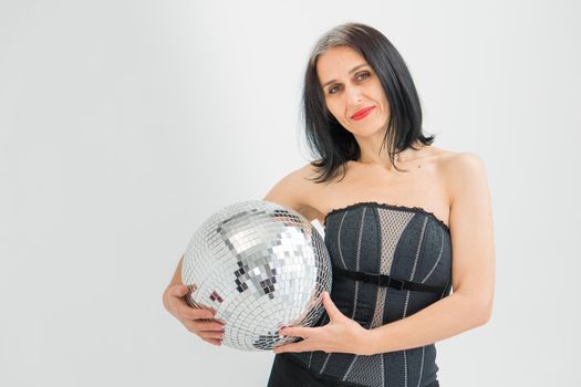 Studio photo of middle aged woman starting getting grey-haired wearing black clothes with silver disco ball in hands on white background, middle age sexy lady, happy life concept.