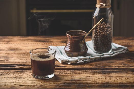 Freshly brewed turkish coffee in drinking glass. Cezve and jar of roasted coffee beans.