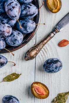 Ripe plums with sliced fruits, leaves and vintage knife over light wooden surface. View from above