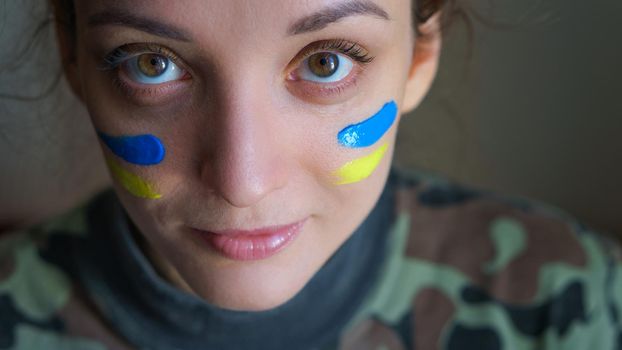 Indoor portrait of young girl with blue and yellow ukrainian flag on her cheek wearing military uniform, mandatory conscription in Ukraine, equality concepts.