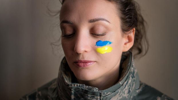 Indoor portrait of young girl with blue and yellow ukrainian flag on her cheek wearing military uniform, mandatory conscription in Ukraine, equality concepts.