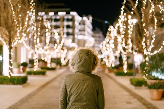 A girl in a hood with fur stands on a street decorated for Christmas in the city. High quality photo