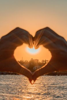 Silhouette against the sunset - hands folded in the shape of a heart. Sun with rays between the palms. Concept for Valentine's Day, declaration of love. Minimalism, space for text, warm colors