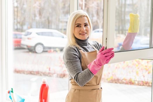 Beautiful young woman is using a duster smiling while cleaning windows in the house.