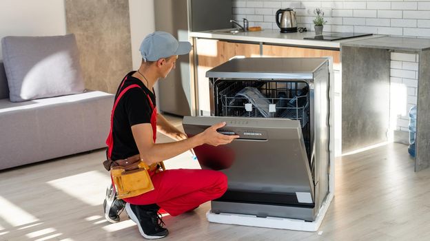 Young Repairman Service Worker Repairing Dishwasher Appliance In Kitchen.