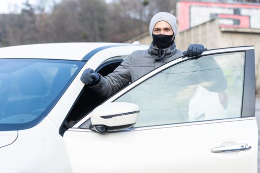 Young adult brunette man in surgical medical mask, jacket posing on city street near the car. Outdoor shot