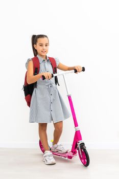 Full length profile shot of a schoolgirl with a backpack riding a scooter isolated on white background.