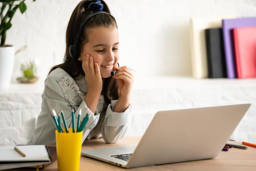 Closeup photo of little pretty pupil school lady talk skype notebook online lesson video call sit desk use earphones distance greet classmates wave arm quarantine study living room indoors