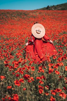 Young woman stands with her back in a long red dress and hat, posing on a large field of red poppies.