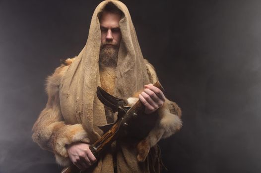 Portrait of a Scandinavian warrior in ancient clothes made of fur and burlap. Stands in the studio surrounded by smoke with an ax in his hands and his eyes closed. ancient muscularity.