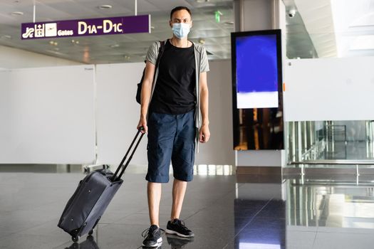 Young European man in gray coat, protective disposable medical mask in airport. Afraid of dangerous N-CoV 2019 influenza coronavirus. backpack, suitcase