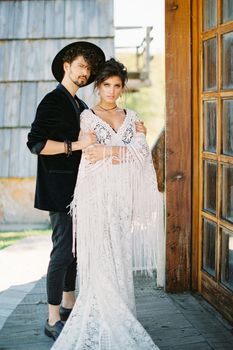 Groom in a hat holds his hands on the shoulders of bride standing on the threshold of a wooden house. High quality photo