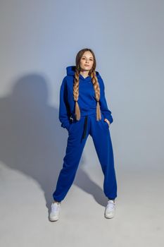 Full length stock photo of stylish pretty Caucasian girl with two long braids wearing bright blue sportive suit and white sneakers. Model in suit holding hands in pockets and smiling at camera.
