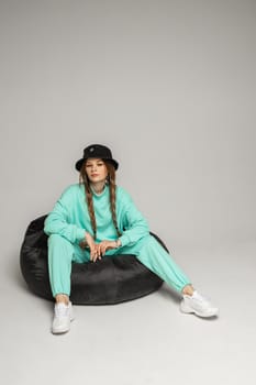 Full length stock photo of trendy brunette with braids wearing black hat and mint sports suit and white sneakers sitting in black bag chair on white background.