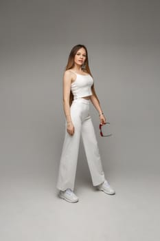 Full length stock photo of attractive brunette in white crop top, jeans and sneakers holding sunglasses in hand and looking at camera. Isolate on white.