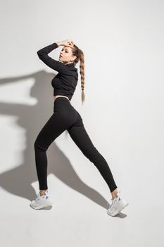 Full length portrait of attractive slender sportive girl in black crop jersey and leggings and white sneakers posing on white background with arms bent towards head.