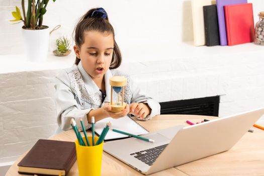 little girl with a laptop at the table and holds an hourglass, waiting for the end of online lessons