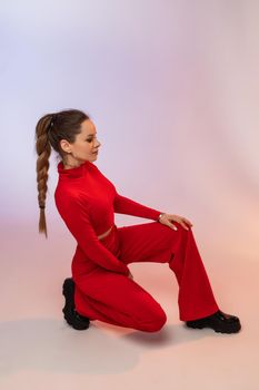 Side view of sitting fashionable model with long braid wearing red suit and black leather boots. Posing on white background.