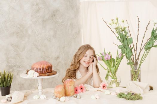 A girl with long hair in a light dress is sitting at the Easter table with cakes, spring flowers and quail eggs. Happy Easter celebration